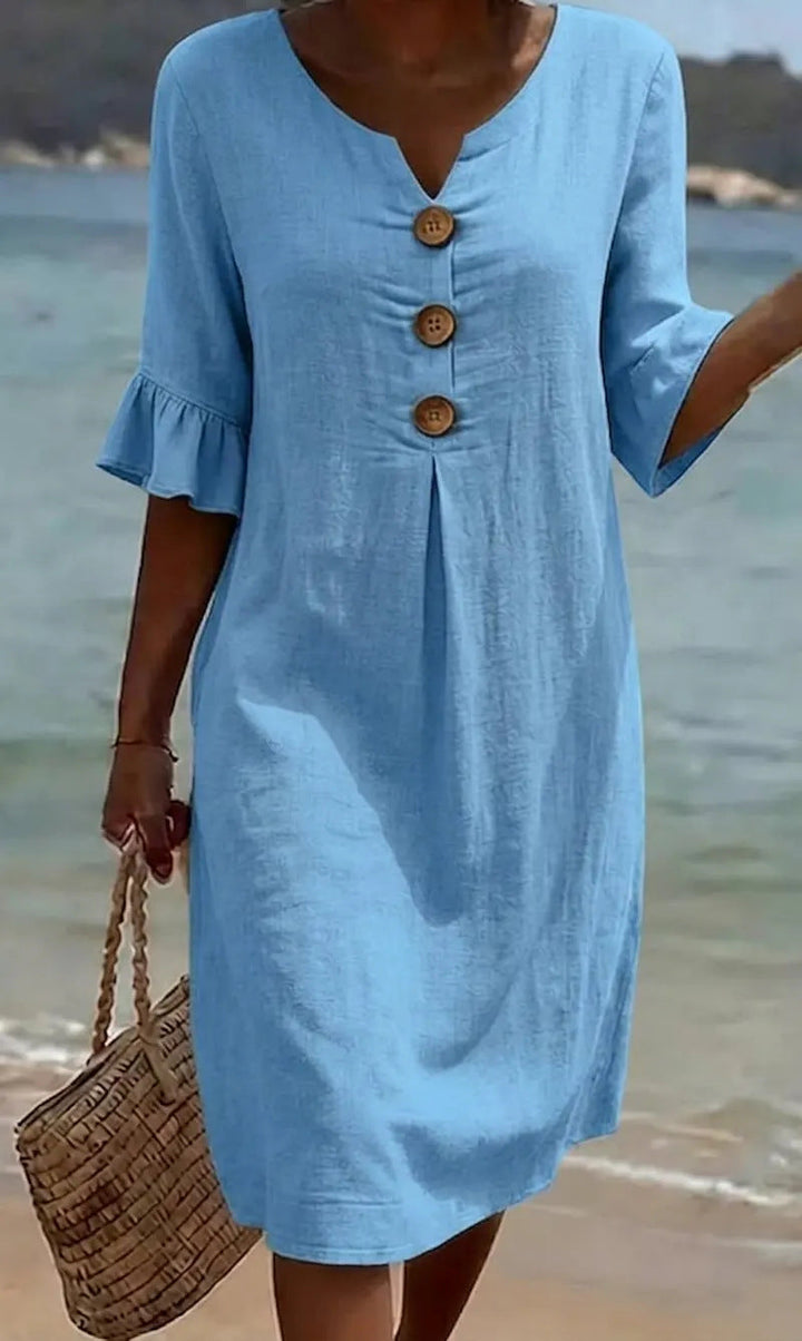 Femme portant une robe en coton lin boutonnée, manches volantées, marchant sur la plage au soleil.